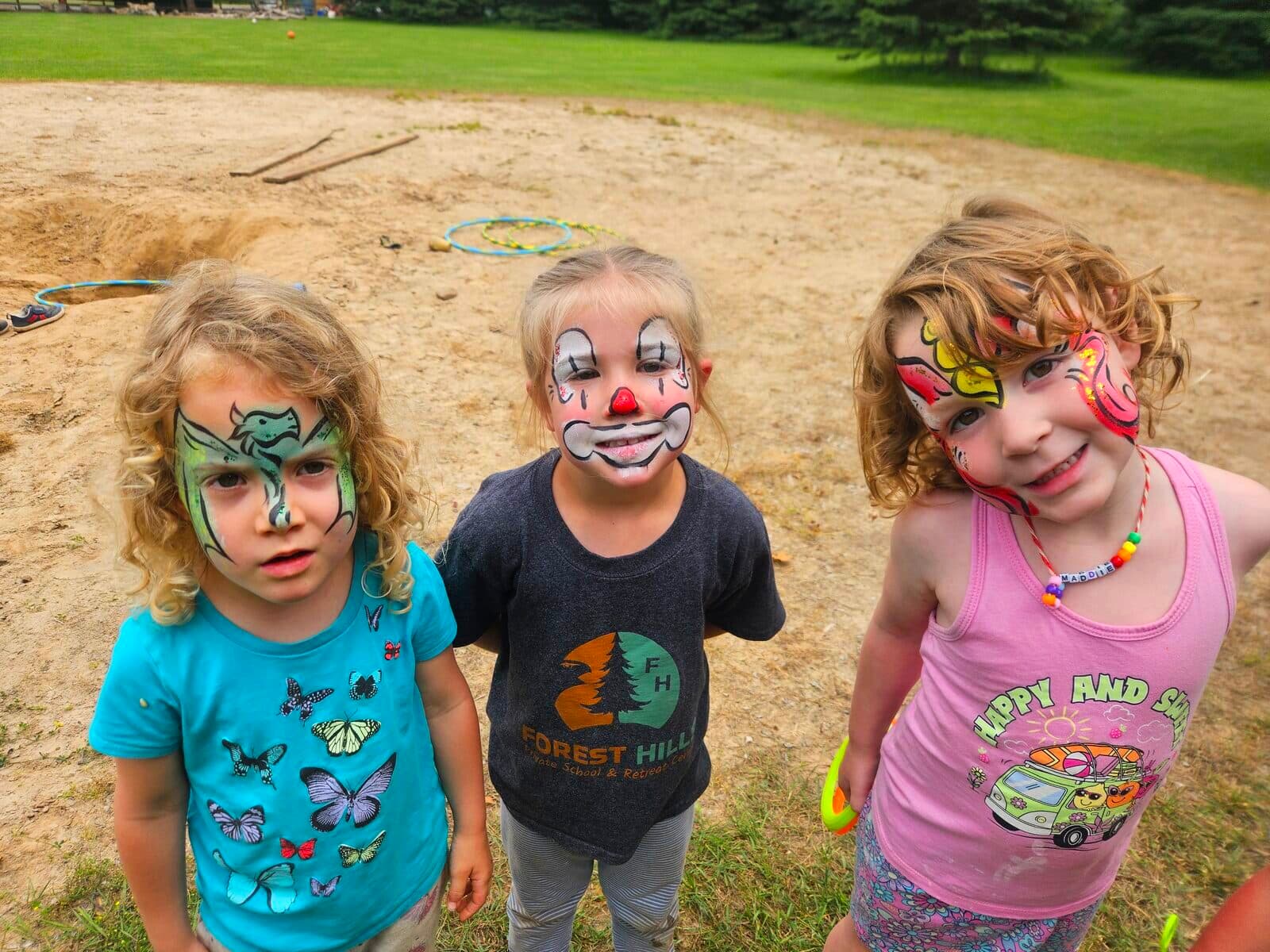 Three kids at a party showing off dragon, clown, and unicorn face paint